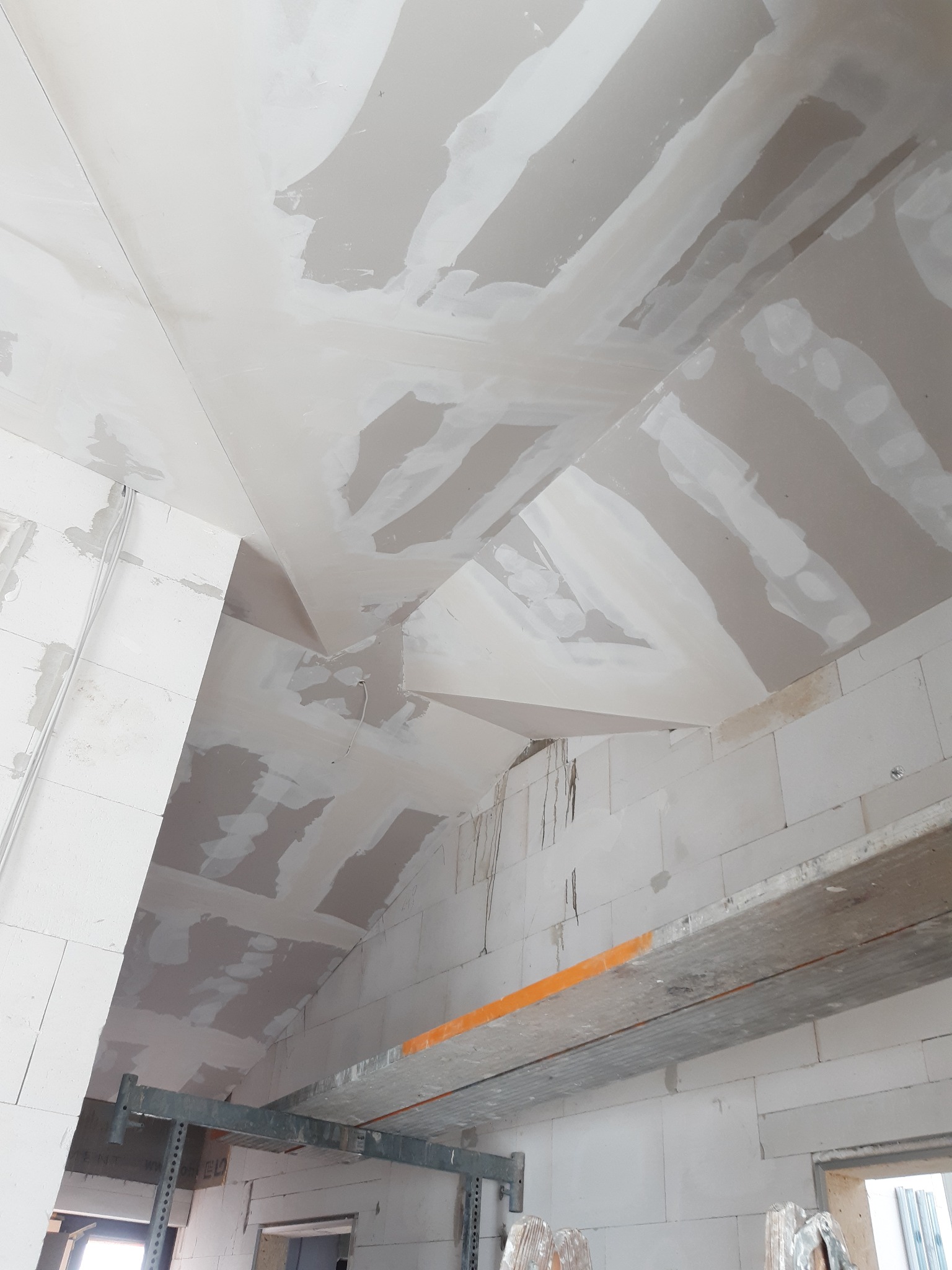 Poddasze z surowymi ścianami i skośnym sufitem z płyt kartonowo-gipsowych w trakcie szpachlowania, widoczne rusztowanie i instalacja elektryczna.