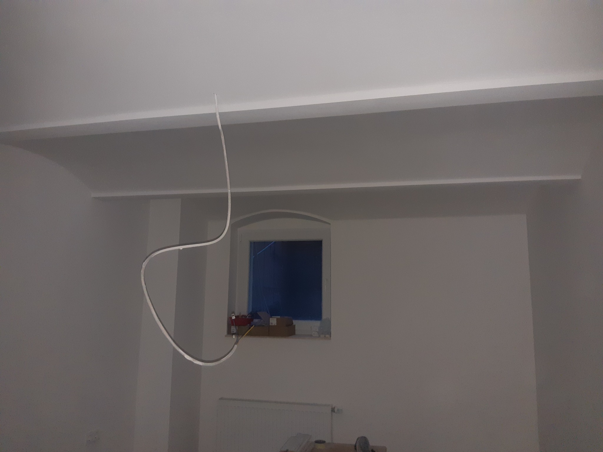 Wnętrze w trakcie prac wykończeniowych: pomalowane ściany na biało, widoczne listwy przysufitowe, okno z zabezpieczeniem, zwisający kabel oświetleniowy i grzejnik pod oknem.