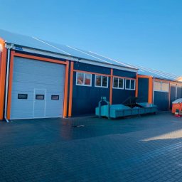 WFB MASZTALERZ SP Z O.O. - Hala przemysłowa z szarą bramą segmentową, obramowaną pomarańczowym kolorem, na tle błękitnego nieba i kostki brukowej.