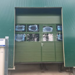 WFB MASZTALERZ SP Z O.O. - Nowa, zielona brama segmentowa z oknami zabezpieczonymi folią ochronną, widoczne nogi montażystów i fragment konstrukcji budynku.