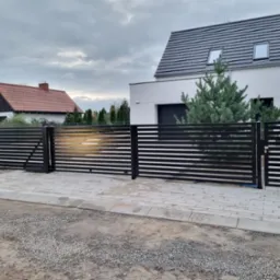 Nowoczesna czarna brama wjazdowa z poziomymi szczeblami, na tle domu z szarą dachówką i świerkiem, ułożona kostka brukowa.