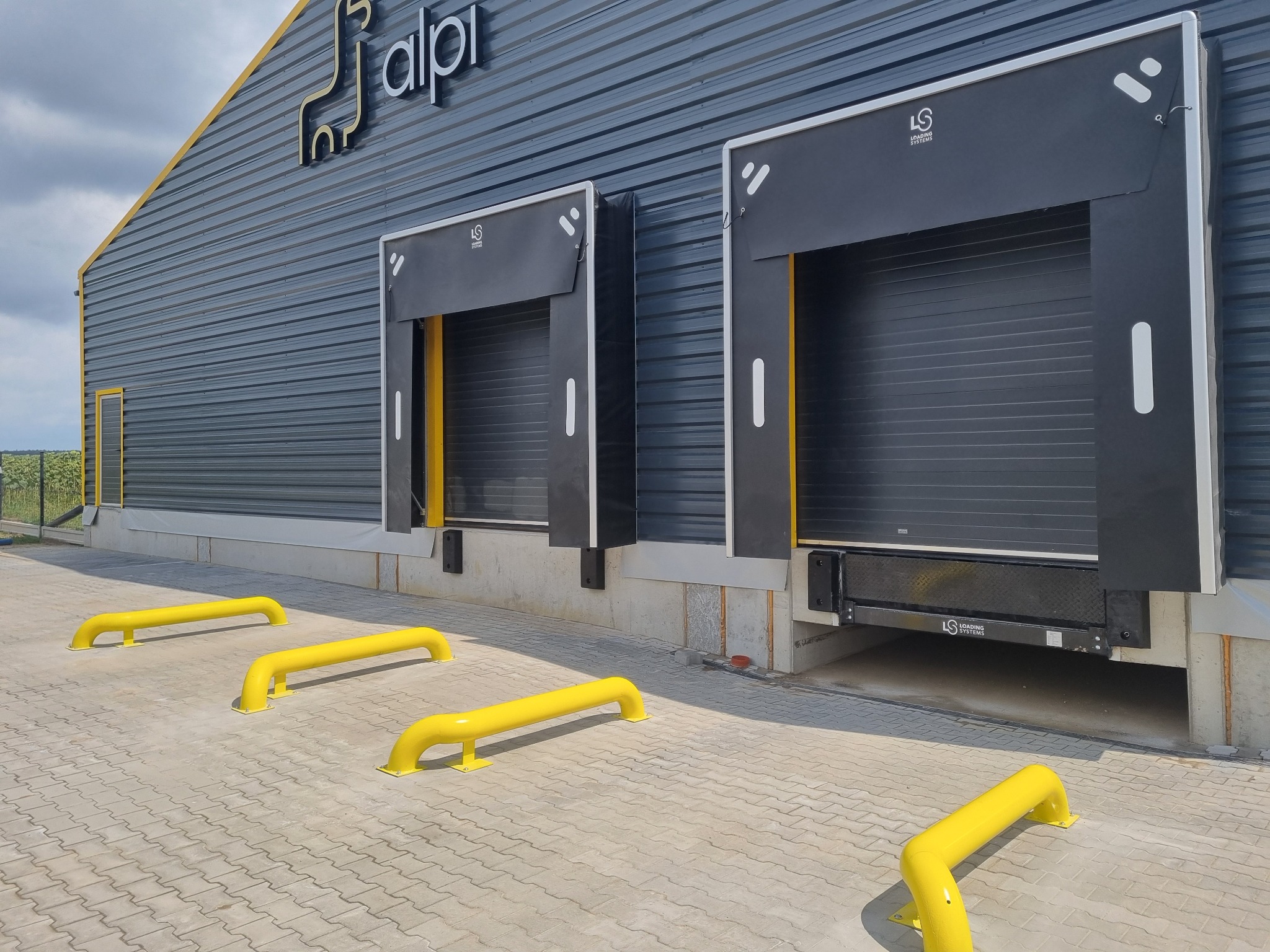 Zewnętrzny widok budynku przemysłowego z dwiema czarnymi bramami segmentowymi z żółtymi akcentami, zabezpieczonymi żółtymi barierami ochronnymi na brukowanym placu.