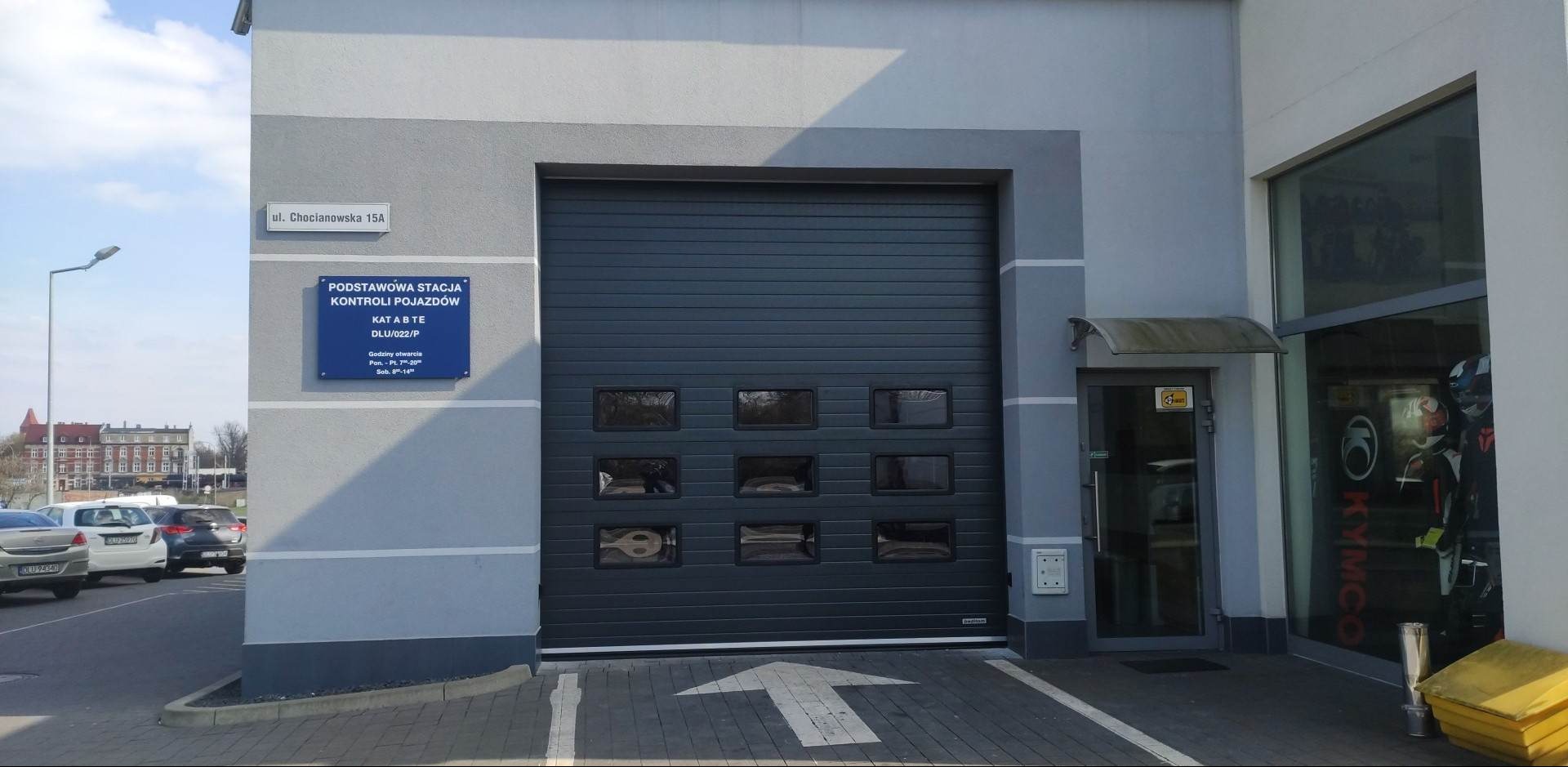 Szara segmentowa brama z sześcioma prostokątnymi okienkami w budynku Podstawowej Stacji Kontroli Pojazdów przy ulicy Chocianowskiej 15A.