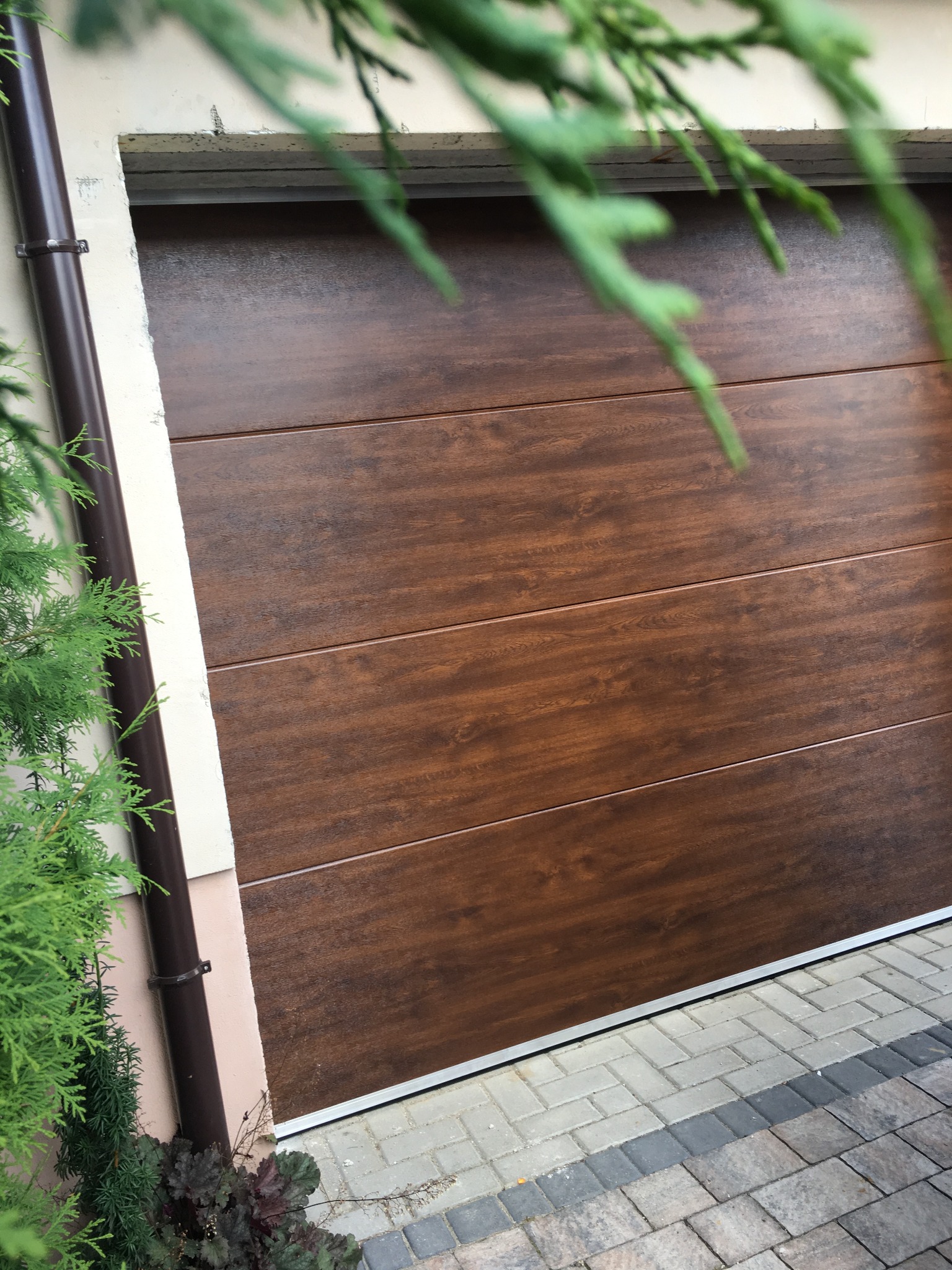 Zamknięta, segmentowa brama garażowa w odcieniach brązu z fakturą drewna, widoczna z boku, z fragmentem elewacji, rynny i roślinności w kadrze.