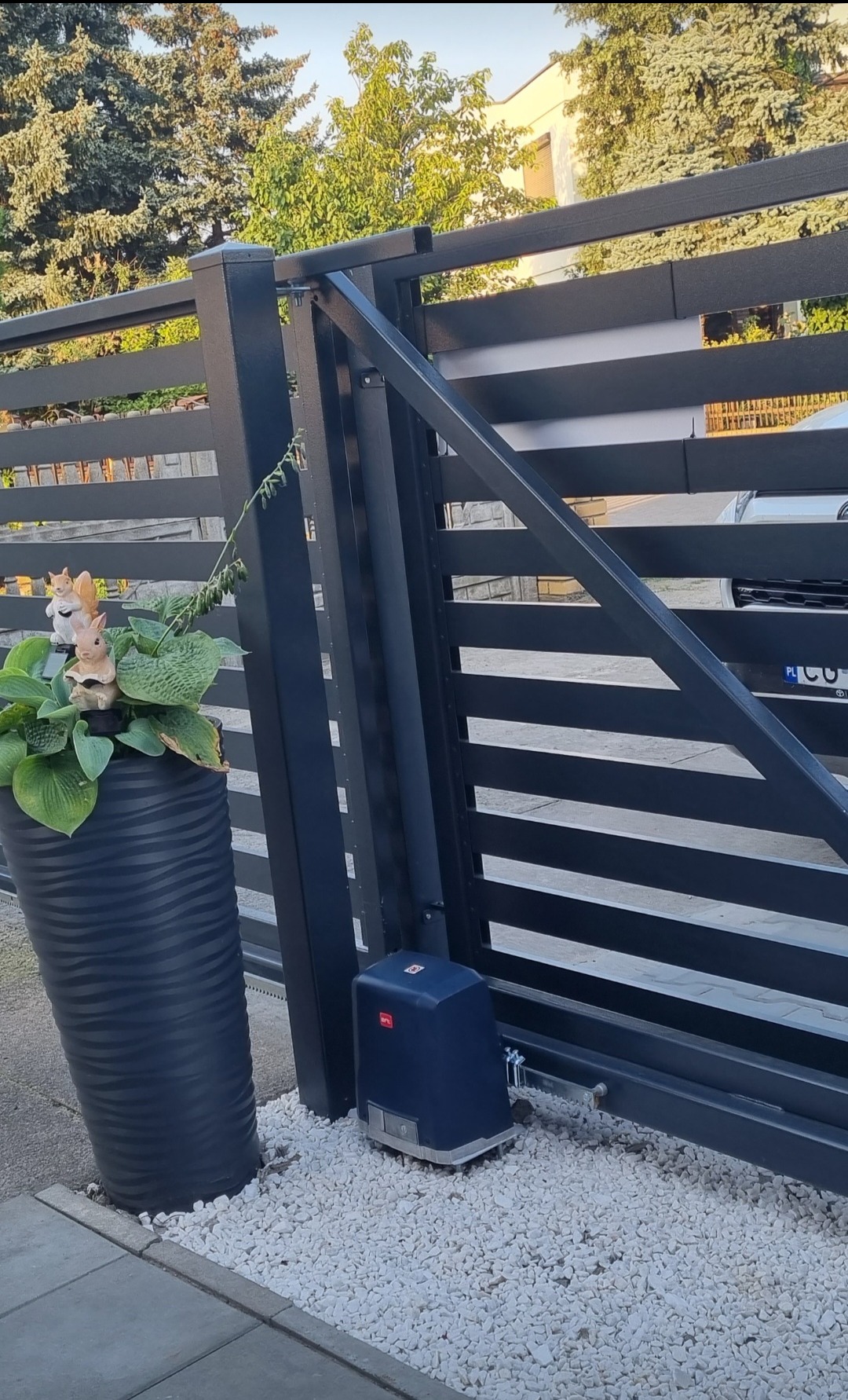 Nowoczesna brama wjazdowa z automatyką BFT, ozdobiona donicą z kwiatami i figurkami wiewiórek, na tle domu i drzew