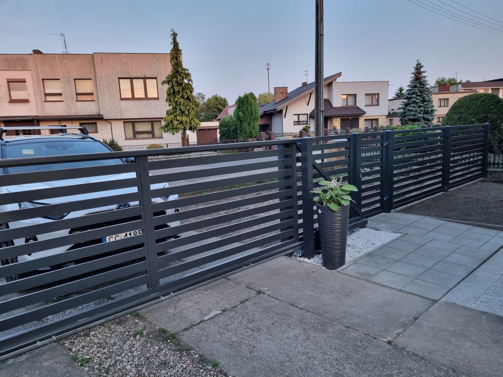 Nowoczesna, przesuwna brama wjazdowa z poziomych, szarych profili, z widocznym napędem i donicą z kwiatami obok, na tle domów jednorodzinnych.