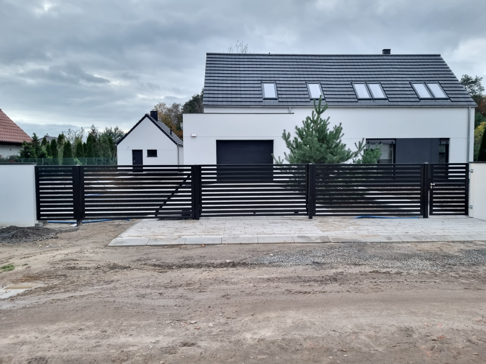 Nowoczesna, pozioma brama wjazdowa z czarnych profili w Zamysłowie, na tle białego domu z szarą dachówką i poddaszem. Teren wokół bramy jeszcze niezagospodarowany.
