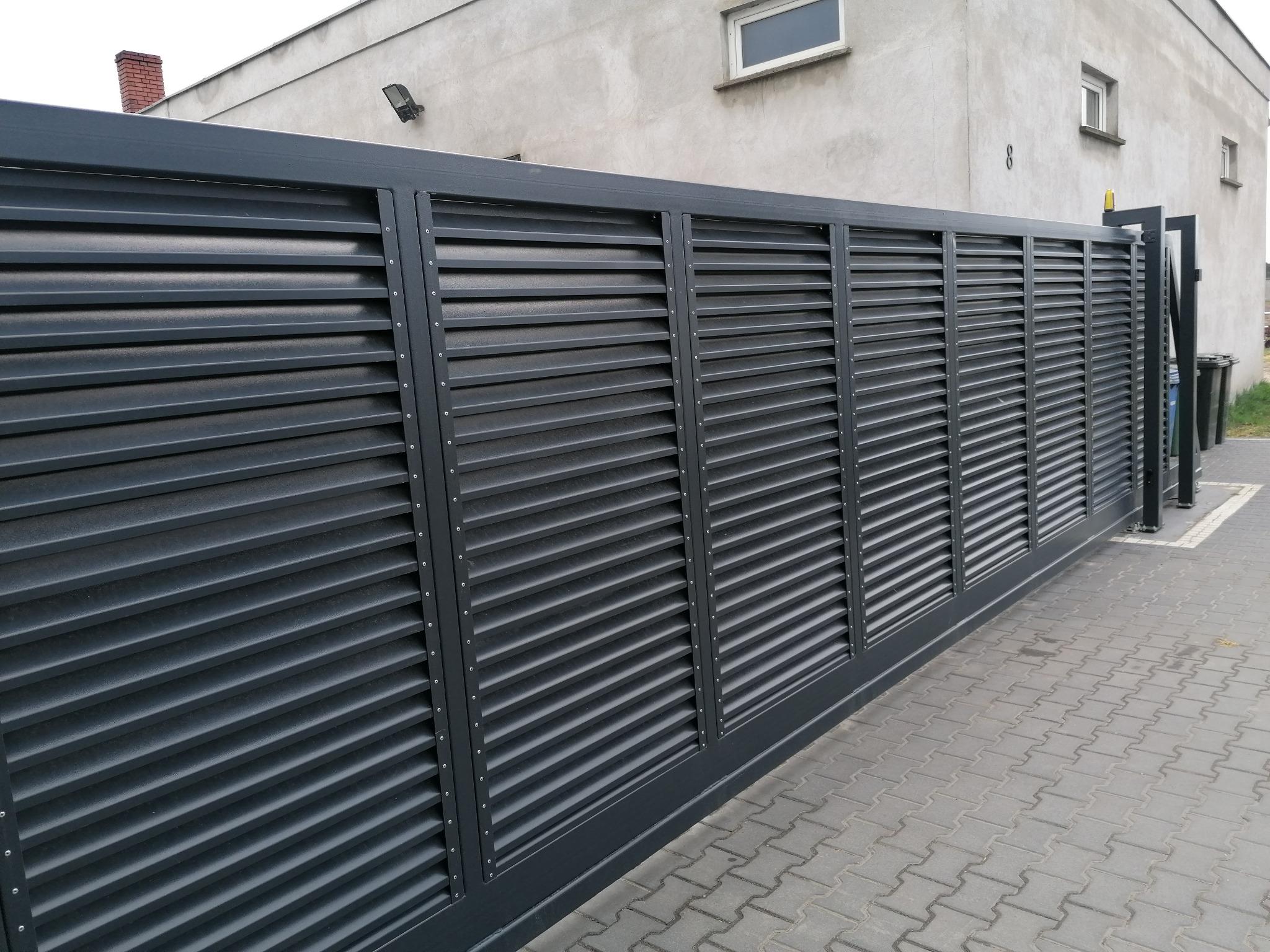 Nowoczesna, przesuwna brama wjazdowa z poziomymi lamelami w kolorze grafitowym, zamontowana przed budynkiem z szarą elewacją i kostką brukową.