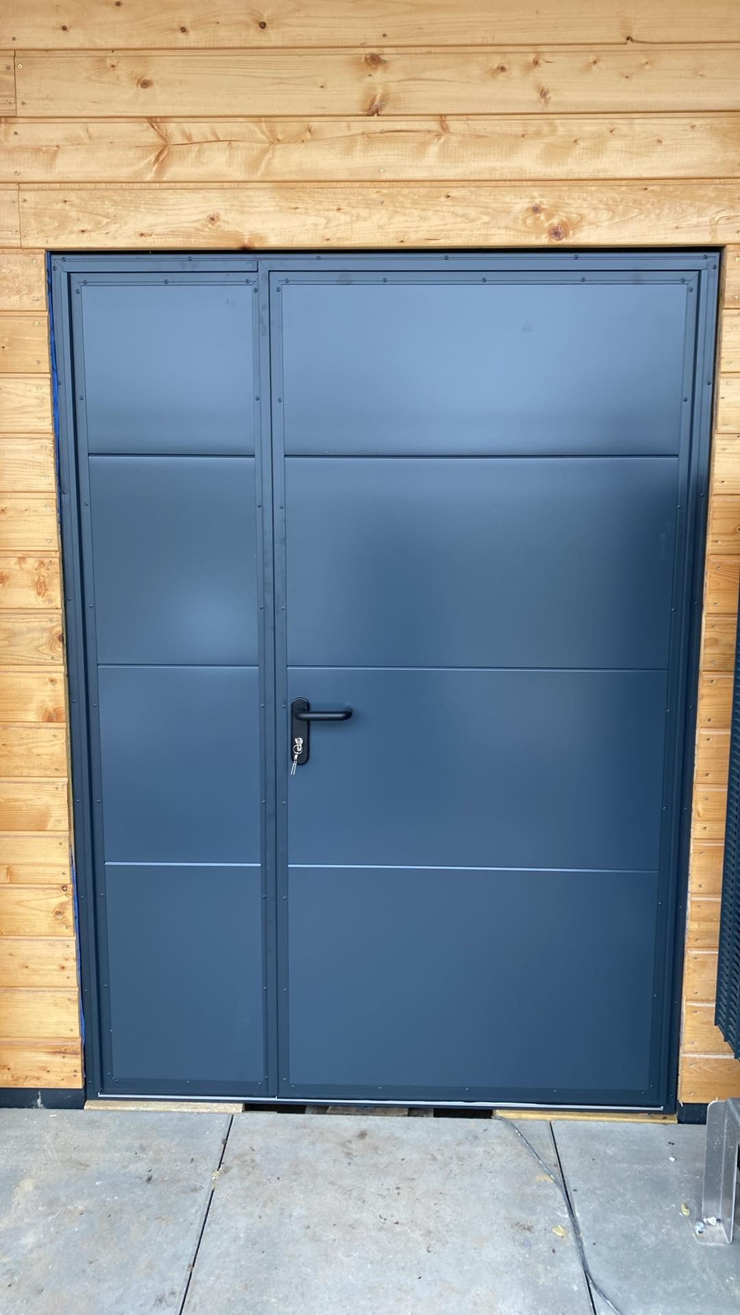 Szara, segmentowa brama garażowa z czarną klamką i kluczem w zamku, zamontowana w drewnianej elewacji budynku.