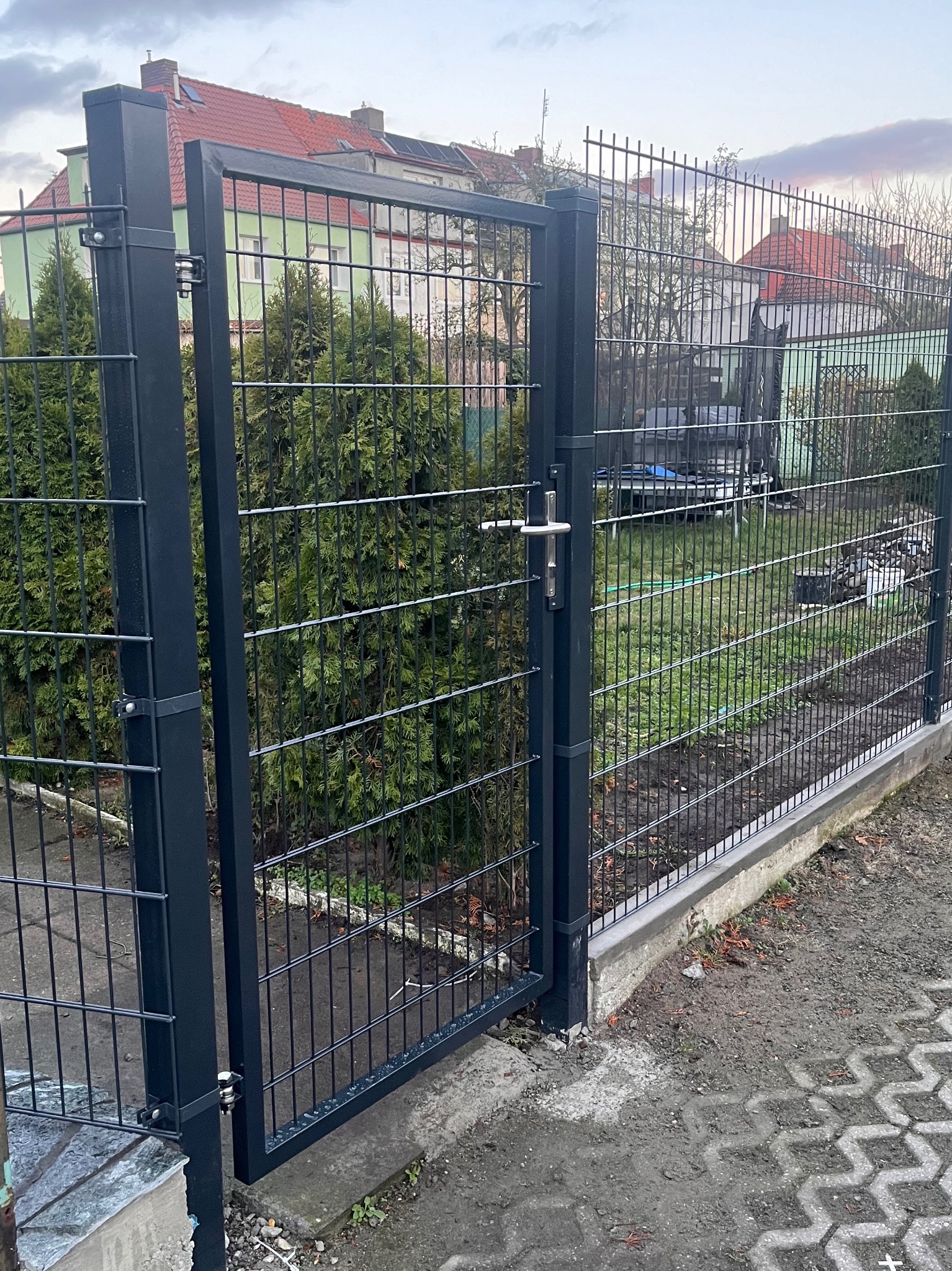 Otwarta, czarna, metalowa furtka ogrodzeniowa z siatki, widok z zewnątrz posesji na trawnik i zabudowania gospodarcze w tle.