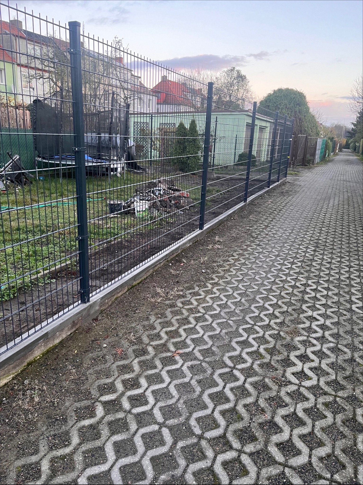 Nowo zainstalowane, ciemne metalowe ogrodzenie panelowe z betonową podmurówką, widok z boku na posesję z kostką brukową, w tle zabudowania mieszkalne i gospodarcze.