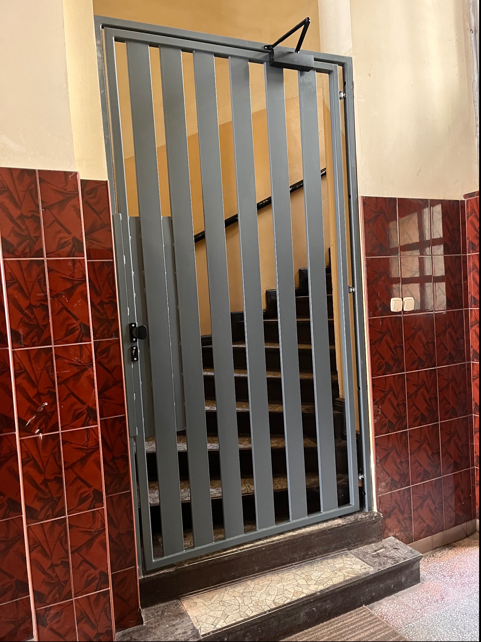 Szara metalowa barierka z pionowych prętów, zabezpieczająca wejście na schody w budynku z czerwono-brązowymi płytkami na ścianach i beżowym tłem, widoczny mechanizm samozamykający na górze.