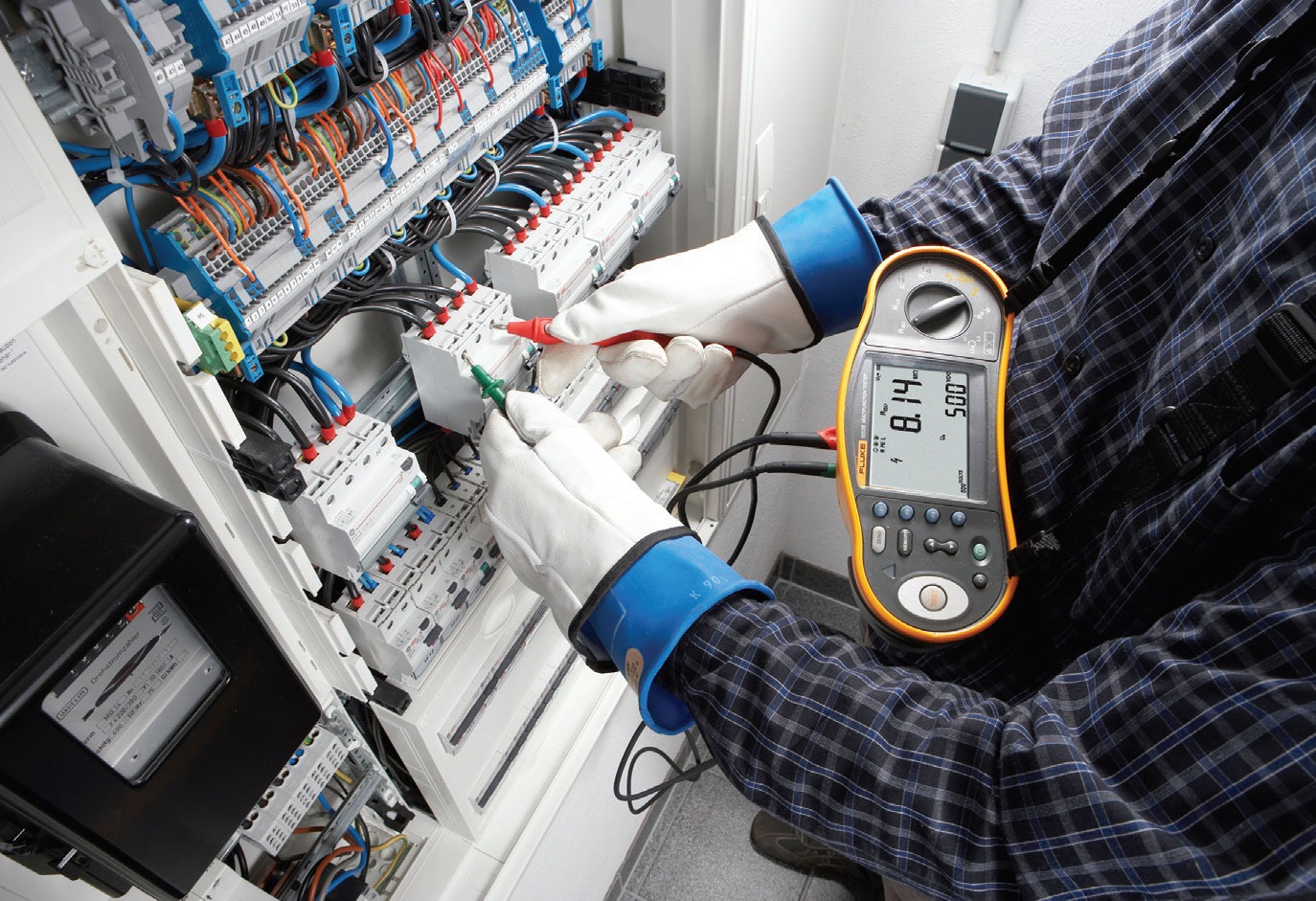 Elektryk w rękawicach ochronnych mierzy parametry w rozdzielnicy elektrycznej za pomocą miernika Fluke, widoczne przewody, bezpieczniki i elementy sterujące.