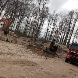 AS-KOMOR PAWEŁ KOMOR - Dwie koparki Atlas i MAN wywożą ziemię na tle lasu bez liści, pracownik w kamizelce odblaskowej nadzoruje prace.