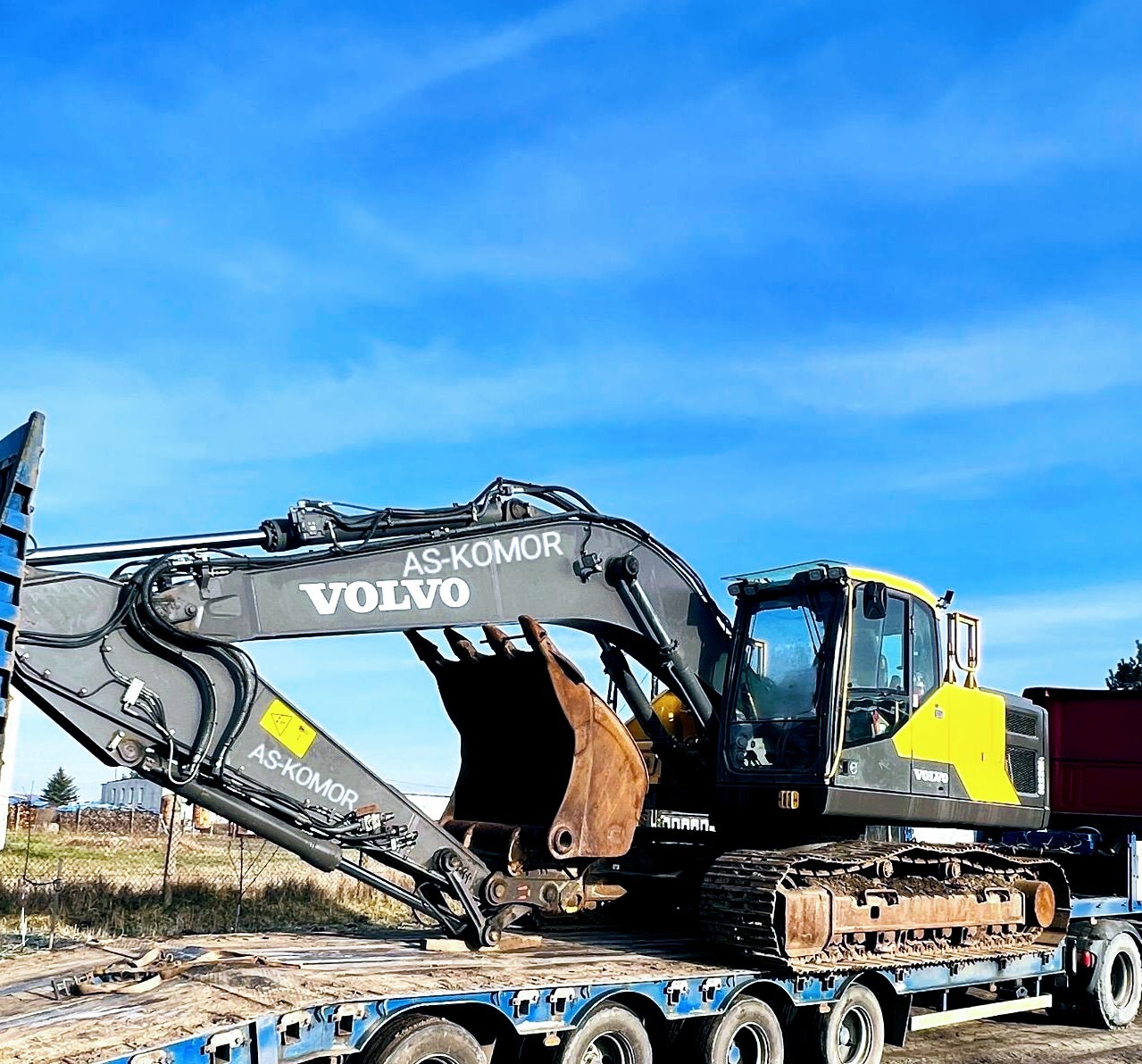 Żółto-szara koparka Volvo z łyżką, z logo AS-KOMOR, przewożona na niskopodwoziowej naczepie, na tle błękitnego nieba.
