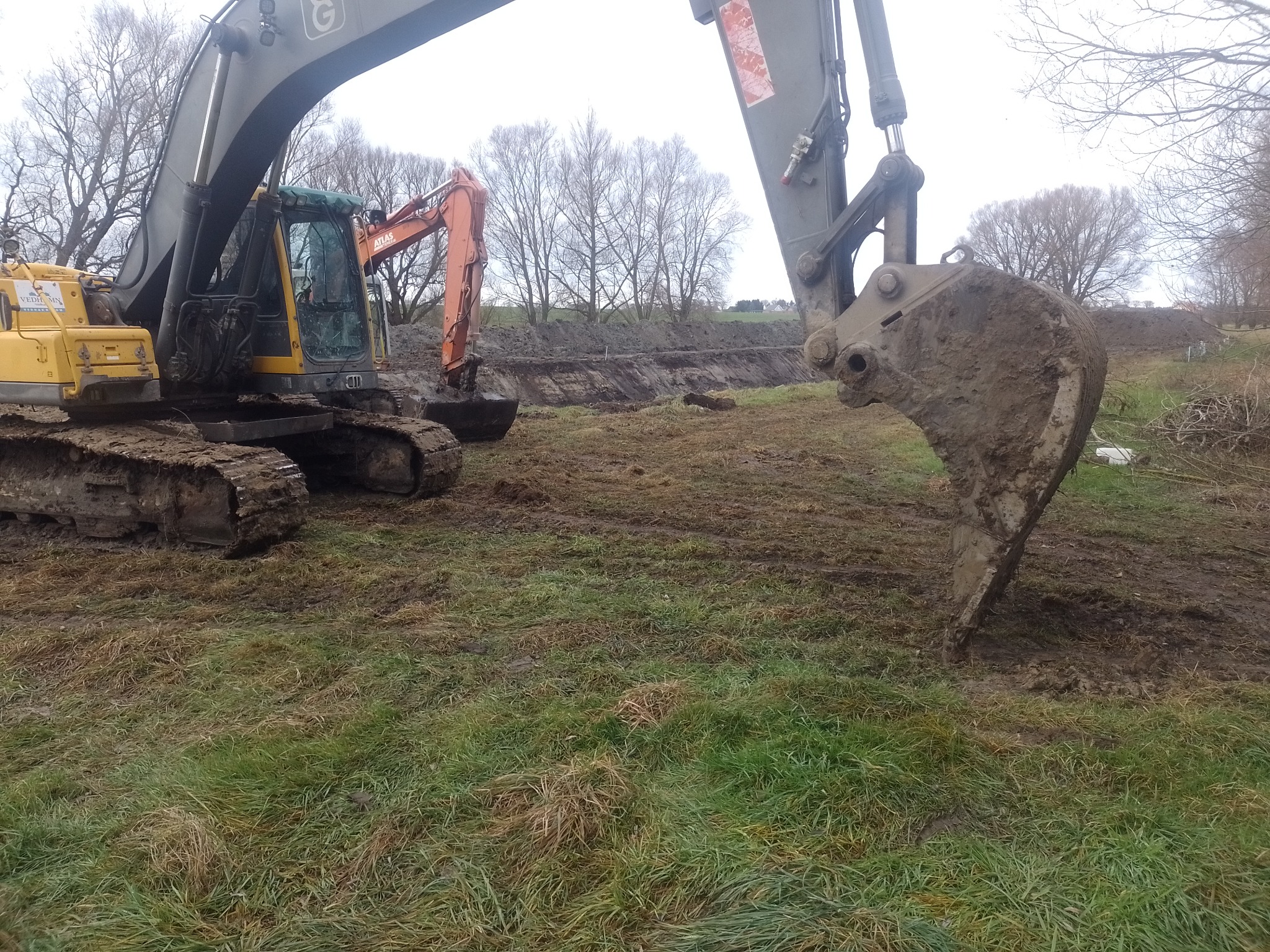 Dwa żółte i pomarańczowe koparki pracują na polu z wykopanym rowem w tle, widoczne błoto na łyżce koparki i na gąsienicach, pochmurne niebo.