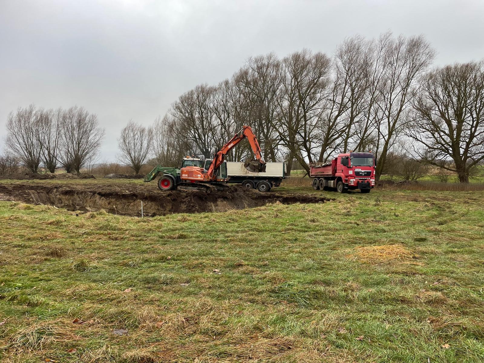 Pomarańczowa koparka nabiera ziemię do skrzyni wywrotki na tle wykopu i czerwonej ciężarówki MAN, widok na łąkę z rzadkimi drzewami pod pochmurnym niebem.