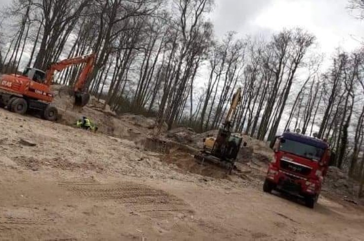 Dwie koparki Atlas i MAN wywożą ziemię na tle lasu bez liści, pracownik w kamizelce odblaskowej nadzoruje prace.