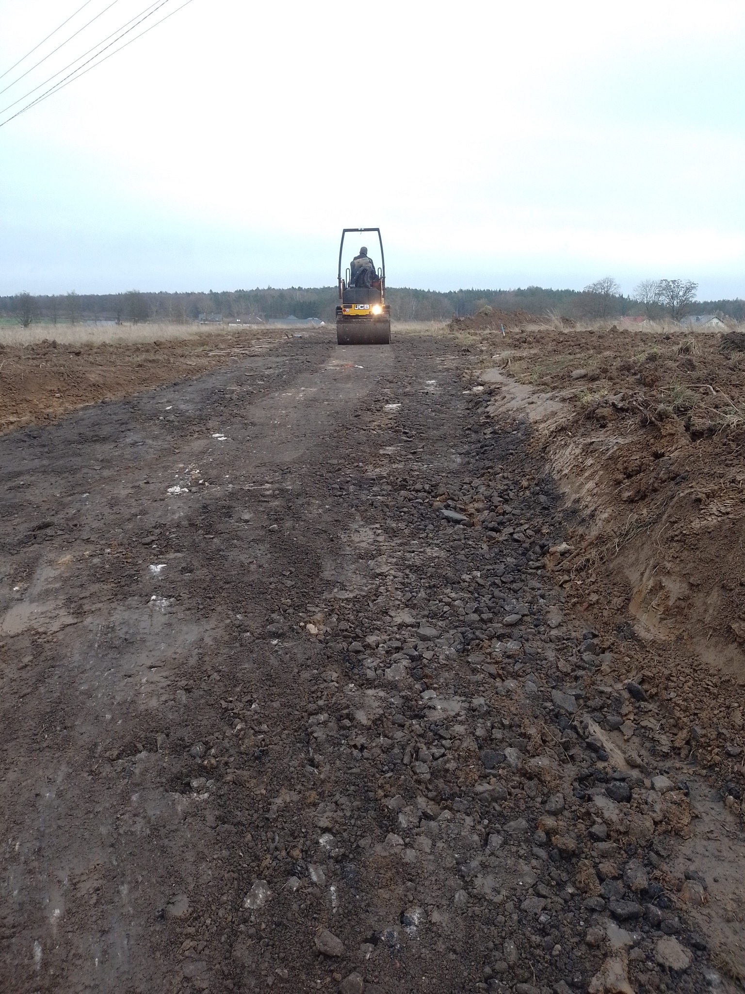 Walec drogowy JCB ubija teren pod budowę drogi gruntowej. Prace ziemne w toku, widoczne nierówności i świeżo usypana ziemia. Pochmurne niebo w tle.