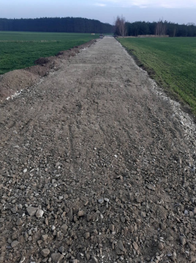 Utwardzona, szutrowa droga polna na tle zielonych pól i lasu. Widoczne ślady opon. Perspektywa z dołu, podkreślająca długość drogi. Lekkie zachmurzenie.