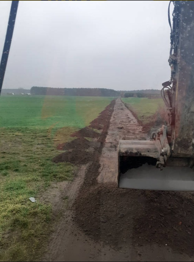 Wykop liniowy w gruncie ornym widziany z kabiny koparki. Świeżo wykopany rów ciągnie się w dal, po prawej fragment maszyny. Pochmurny dzień.