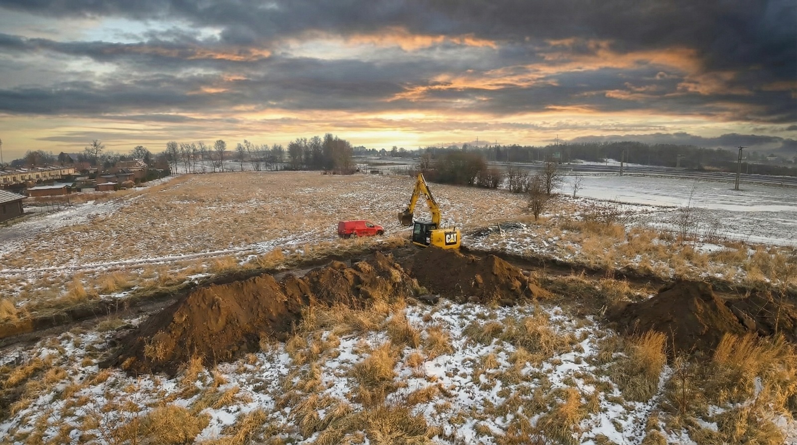 Żółta koparka CAT w trakcie prac ziemnych na polu z resztkami śniegu, widoczny wykop i sterta ziemi, w tle czerwony bus i zachmurzone niebo o zachodzie słońca.