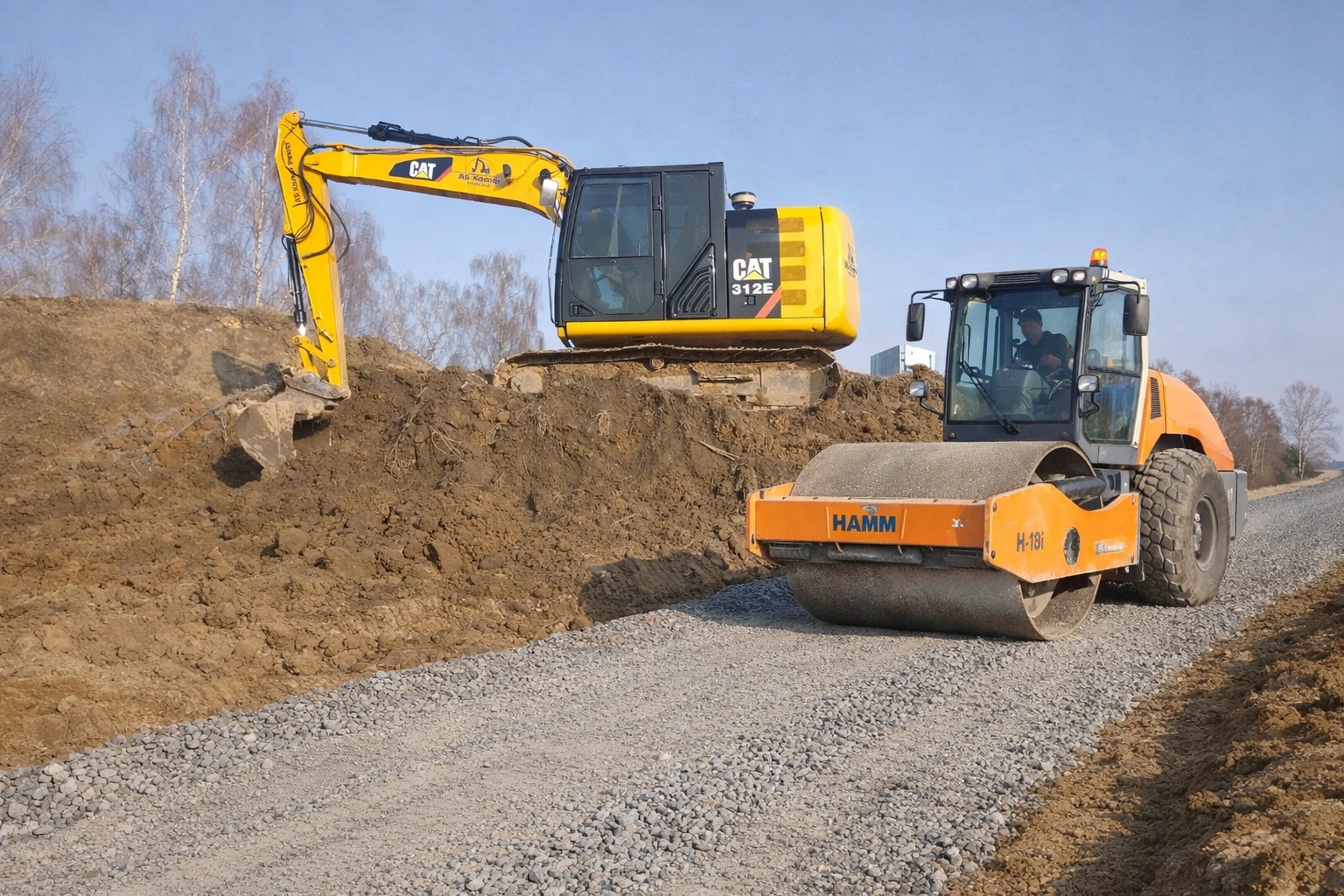Żółta koparka CAT 312E i pomarańczowy walec HAMM H-19I na żwirowej drodze w trakcie budowy, z widocznym operatorem w walcu i wykopem ziemnym w tle.