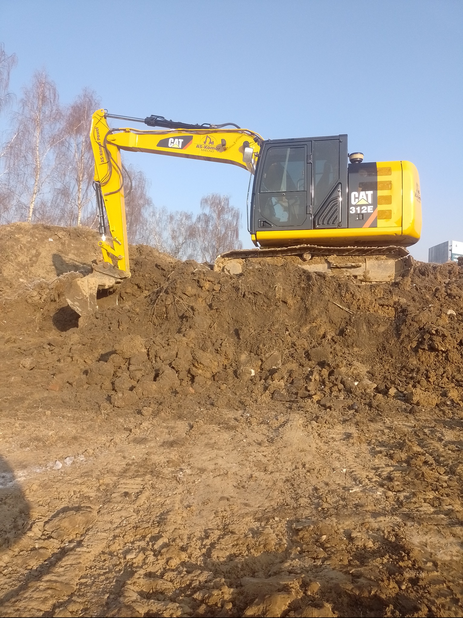 Żółta koparka CAT 312E na wzniesieniu z ziemi, w tle drzewa i błękitne niebo. Widoczny wysięgnik z łyżką zagłębiający się w ziemię. Praca w toku.