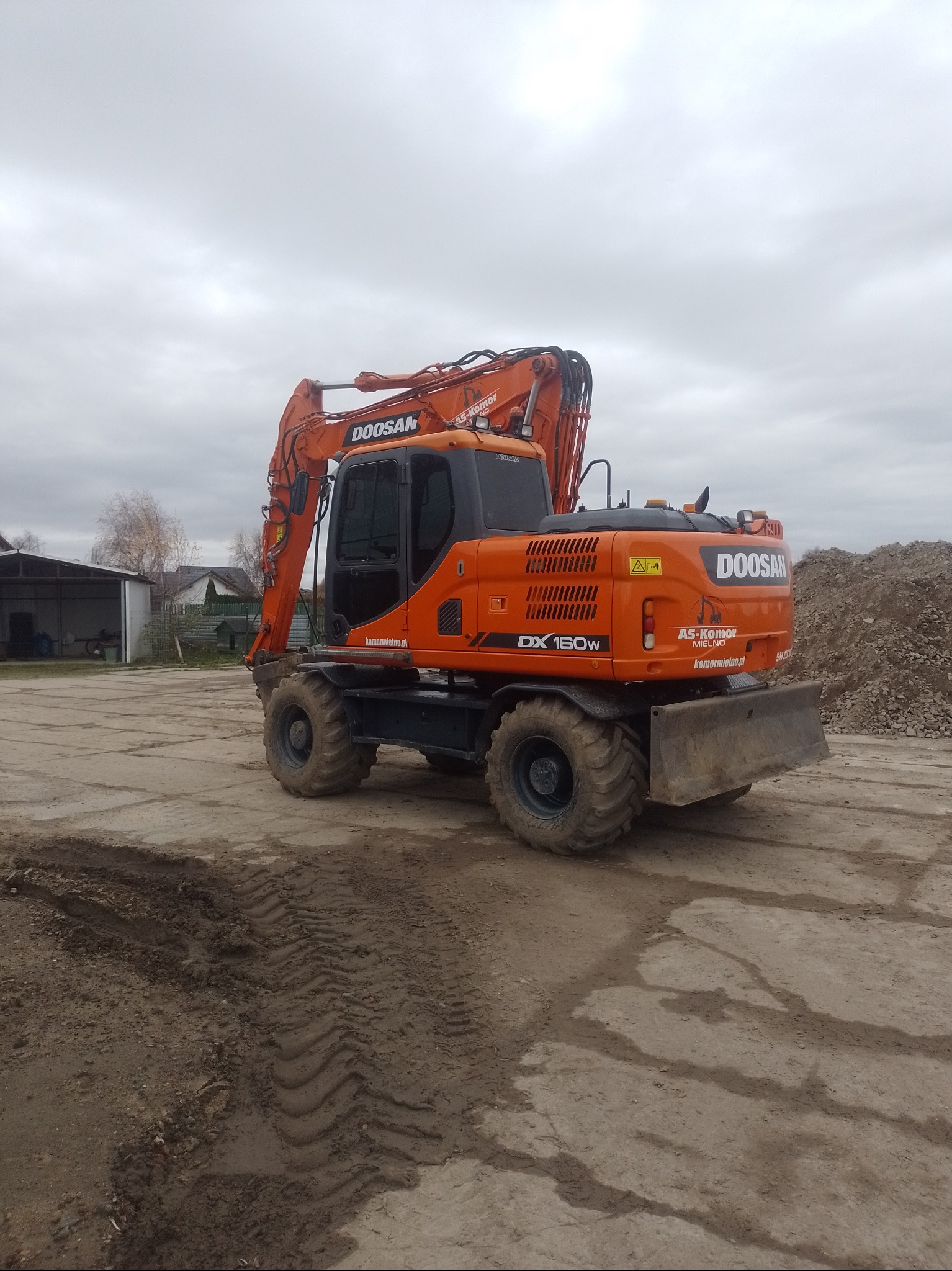 Pomarańczowa koparka kołowa Doosan DX160W na placu budowy, z widocznymi śladami opon na ziemi i stertą gruzu w tle. Pochmurne niebo.