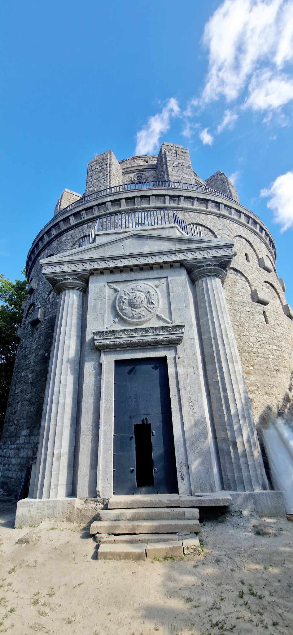 Monumentalna, kamienna wieża z kolumnami i zdobieniami, zwieńczona platformą widokową, pod błękitnym niebem.