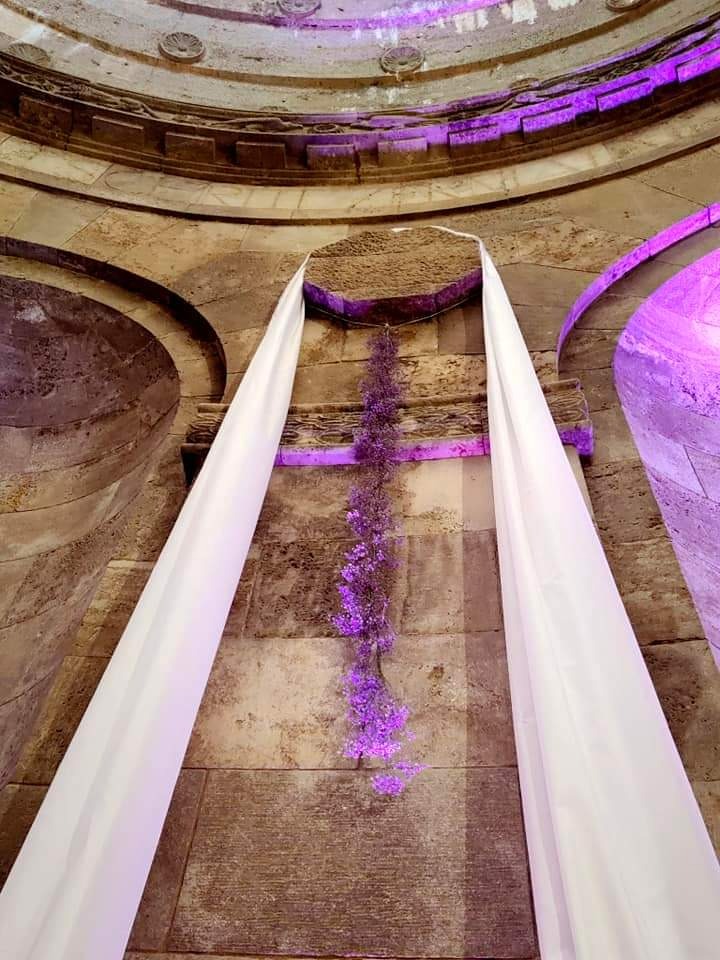 Dekoracja ślubna w zabytkowej rotundzie: białe draperie i kaskada drobnych fioletowych kwiatów na tle kamiennej ściany, oświetlenie w kolorze fioletowym.