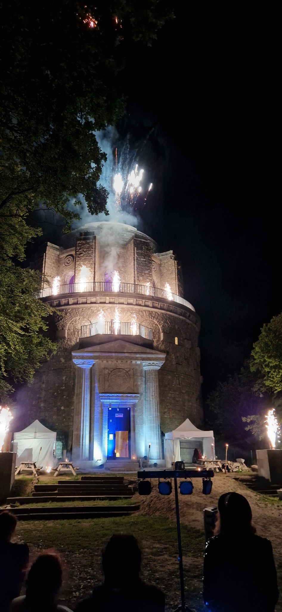 Nocna scena plenerowa z iluminowanym budynkiem rotundy, efekty pirotechniczne nad budowlą i wokół niego, w tle drzewa i publiczność.