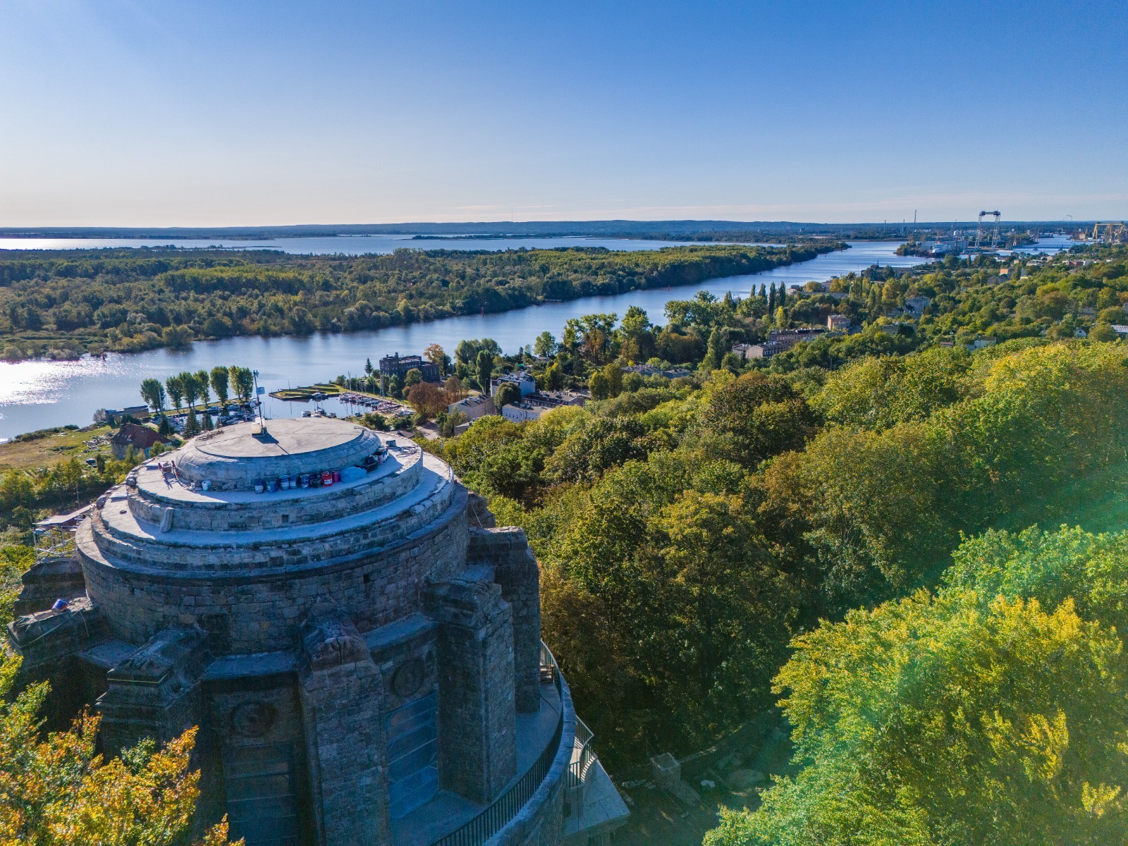 Widok z lotu ptaka na odrestaurowaną kamienną wieżę widokową z tarasem, otoczoną zielenią, z panoramą rzeki i miasta w tle.