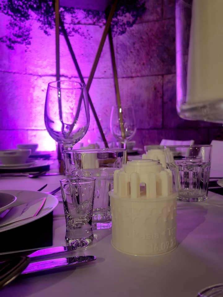 Ustawiony stół z kieliszkami i dekoracyjnym, białym elementem z napisem oświetlony fioletowym światłem w tle.