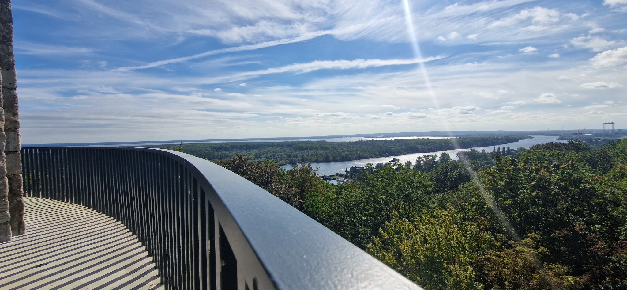 Widok z platformy widokowej na rzekę, las i miasto w oddali, z widoczną metalową barierką i fragmentem kamiennej konstrukcji.