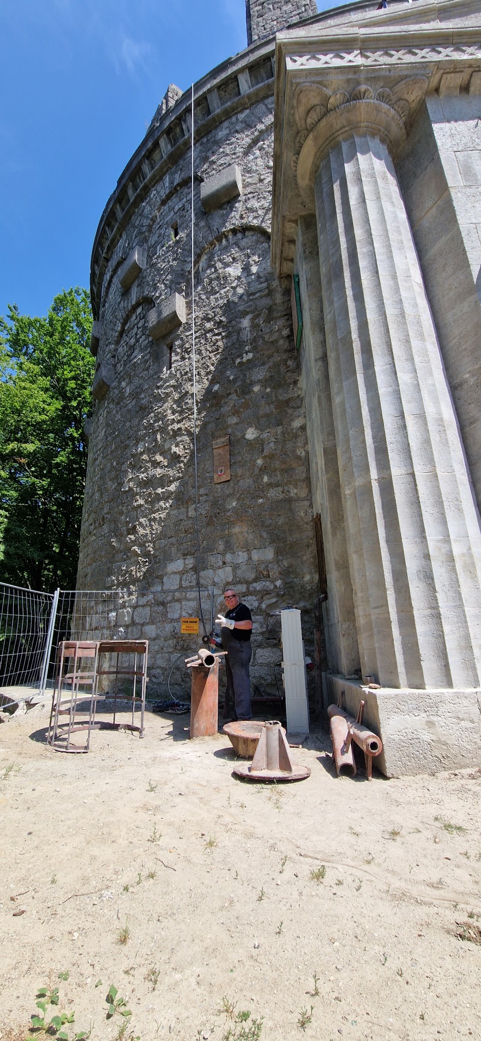 Pracownik w okularach ochronnych szlifuje metalowy element u podnóża kamiennej wieży z kolumną, prawdopodobnie podczas renowacji zabytku.