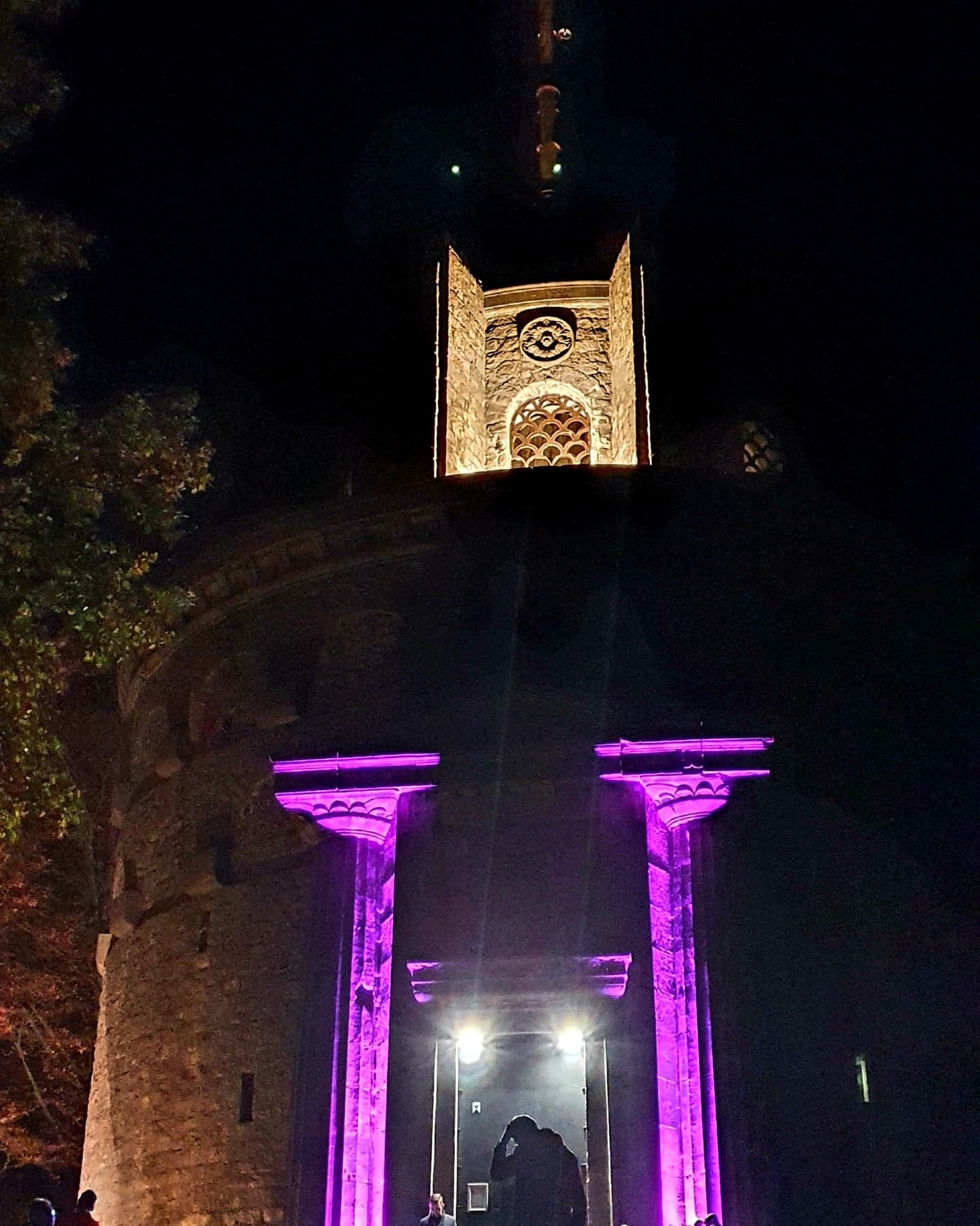 Nocne oświetlenie wieży zamkowej z zegarem, podświetlonej fioletowym światłem, widzianej z dołu.