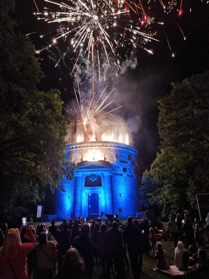 Nocny pokaz sztucznych ogni nad podświetloną na niebiesko rotundą, tłum obserwatorów w tle.