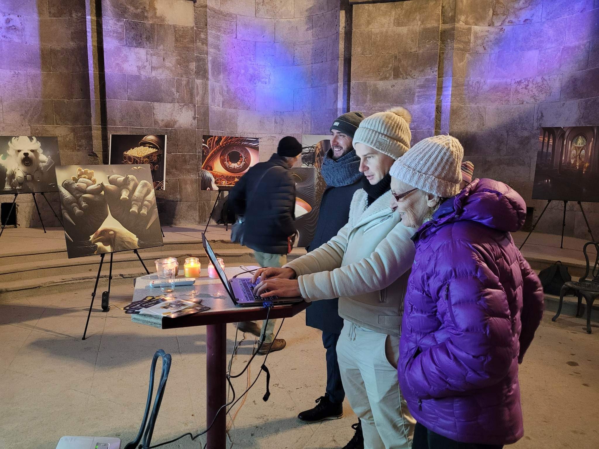 Wystawa fotografii w kamiennej sali z fioletowym oświetleniem; osoby oglądają zdjęcia na sztalugach, jedna osoba pracuje na laptopie przy stole, obok świece i materiały reklamowe.