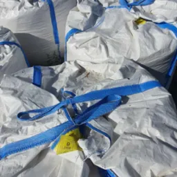 Stos białych, worków big-bag z niebieskimi pasami, wypełnionych materiałem sypkim, leżących na zewnątrz, z widoczną żółtą etykietą na jednym z worków.
