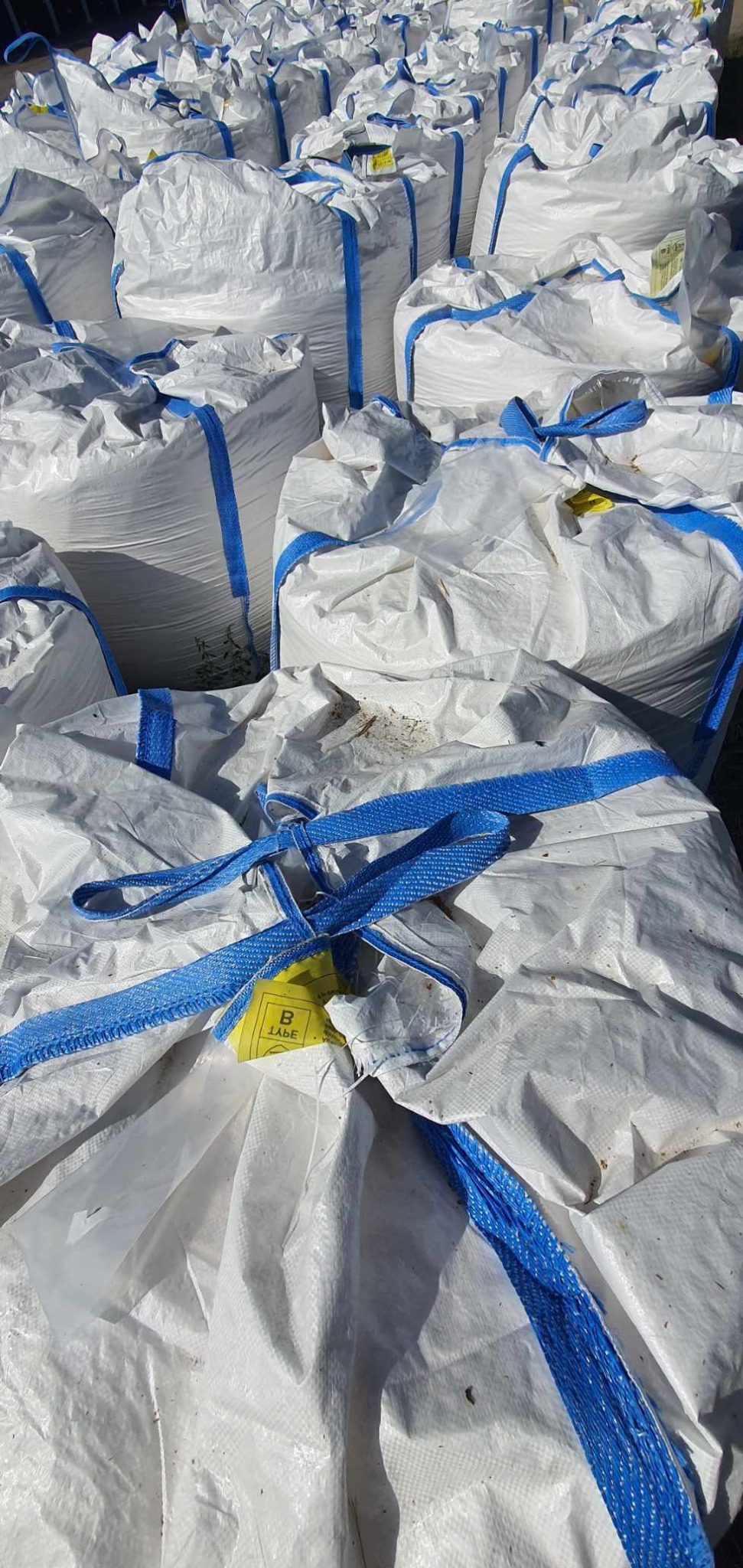 Stos białych, worków big-bag z niebieskimi pasami, wypełnionych materiałem sypkim, leżących na zewnątrz, z widoczną żółtą etykietą na jednym z worków.