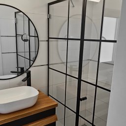 SEAMLESS WALLS CONSTRUCTION SPÓŁKA Z OGRANICZONĄ ODPOWIEDZIALNOŚCIĄ - Nowoczesna łazienka z kabiną prysznicową ze szklanymi drzwiami z czarnymi profilami, okrągłe lustro z czarną ramą i umywalka nablatowa na drewnianej szafce.