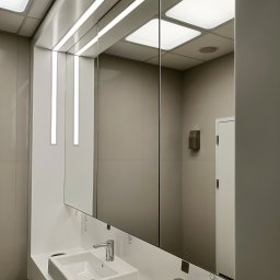 SEAMLESS WALLS CONSTRUCTION SPÓŁKA Z OGRANICZONĄ ODPOWIEDZIALNOŚCIĄ - Nowoczesna łazienka biurowa z dwiema białymi umywalkami, dużym lustrem z podświetleniem LED i dozownikiem mydła na ścianie z szarych płytek.