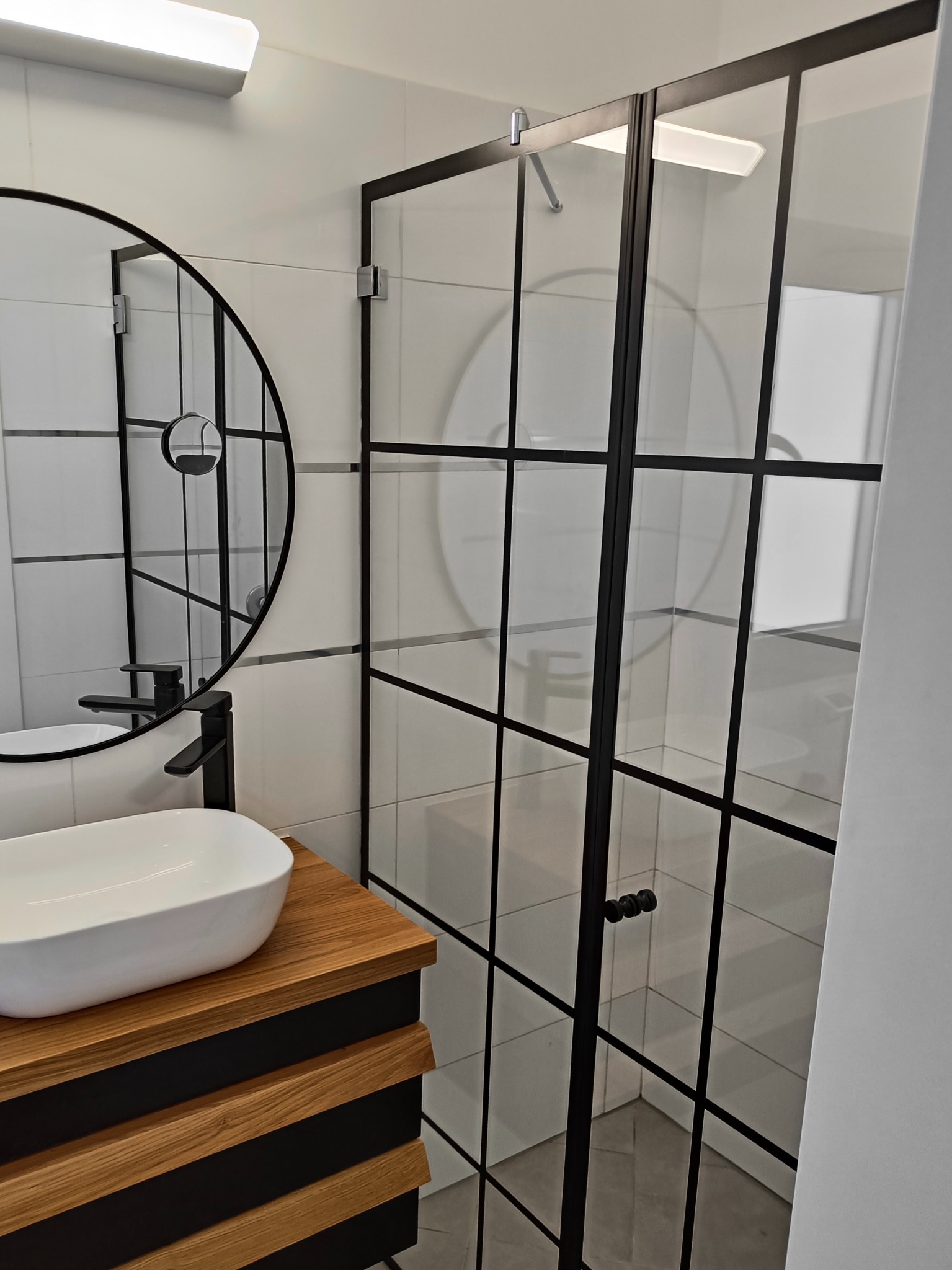 Nowoczesna łazienka z kabiną prysznicową ze szklanymi drzwiami z czarnymi profilami, okrągłe lustro z czarną ramą i umywalka nablatowa na drewnianej szafce.