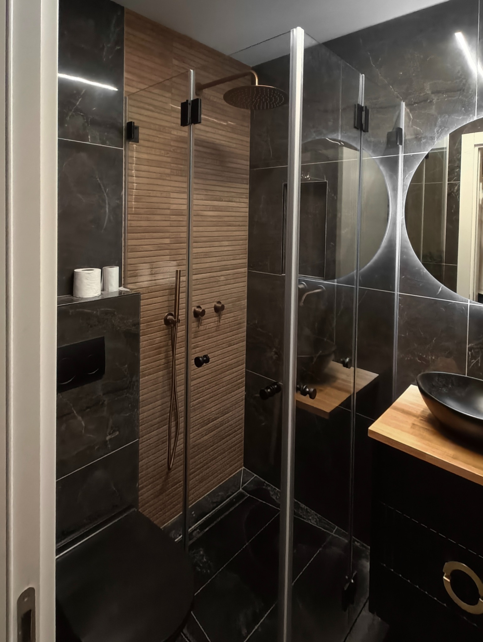 Nowoczesna łazienka z kabiną prysznicową ze szkła hartowanego, armaturą w kolorze brązu i czarnymi płytkami imitującymi marmur.