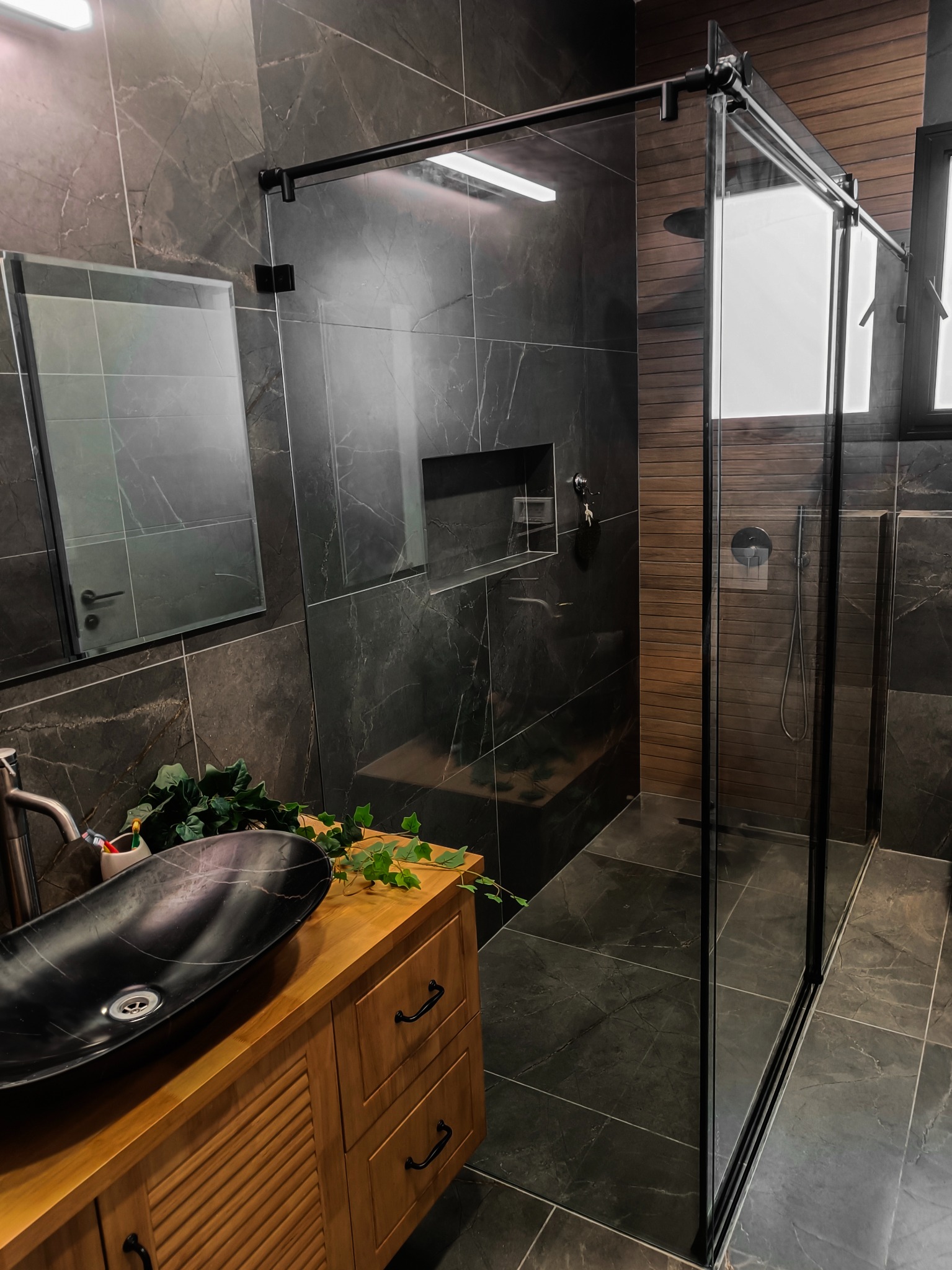 Nowoczesna łazienka z kabiną prysznicową ze szklanymi drzwiami przesuwnymi, czarną umywalką nablatową na drewnianej szafce i marmurowymi płytkami na ścianach.