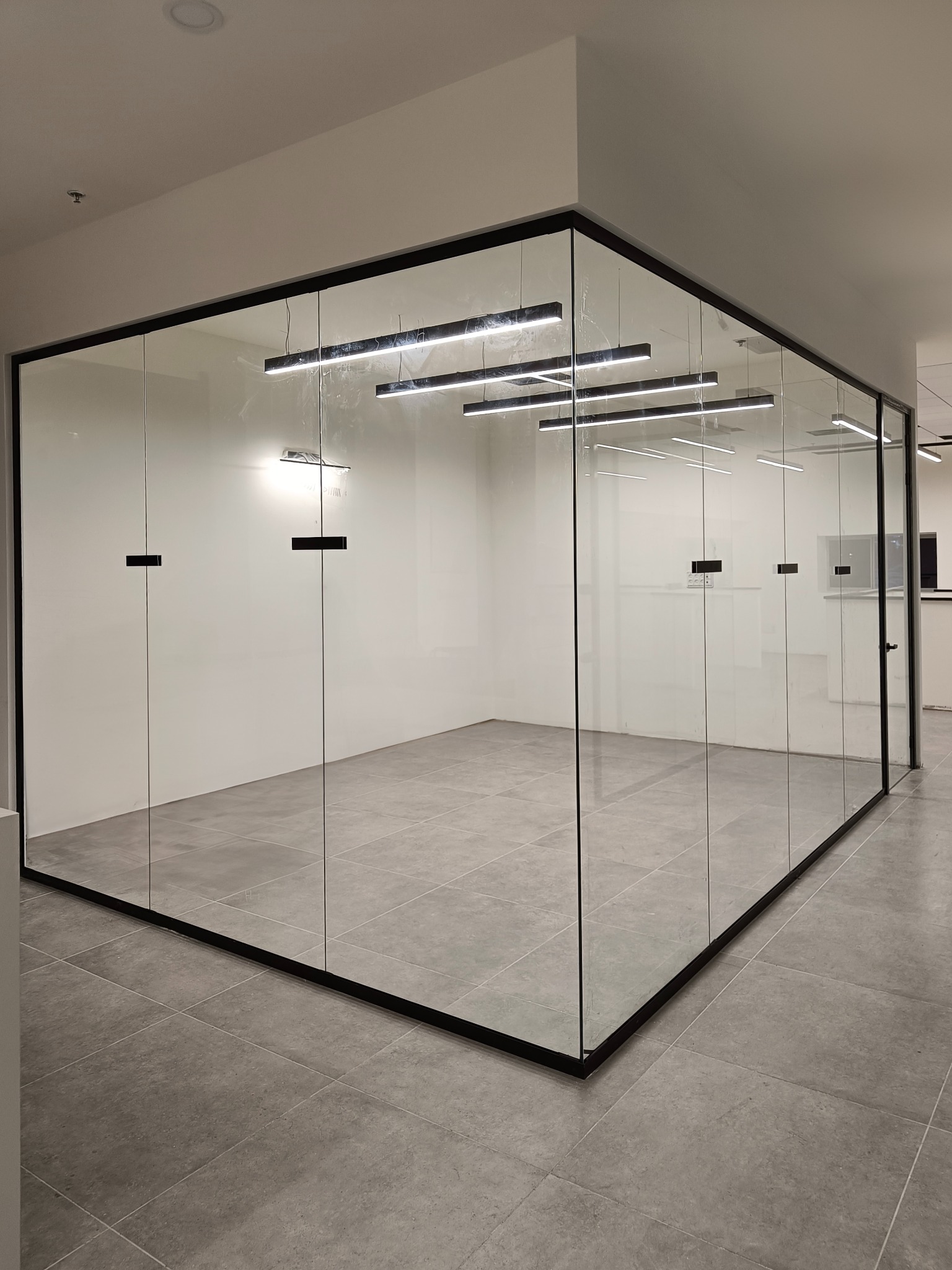 Szklana ścianka działowa z czarnymi ramami w biurze, widoczne minimalistyczne uchwyty na drzwiach i nowoczesne oświetlenie sufitowe.