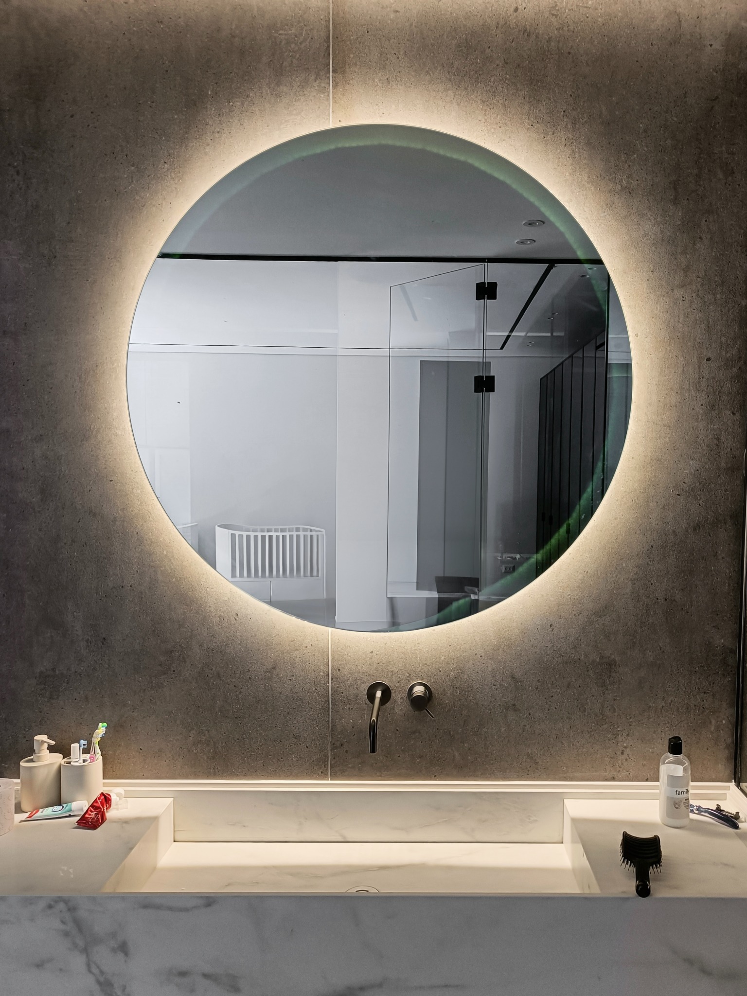 Nowoczesna łazienka z okrągłym lustrem LED, odbijającym szklane drzwi kabiny prysznicowej i białe łóżeczko dziecięce w tle.