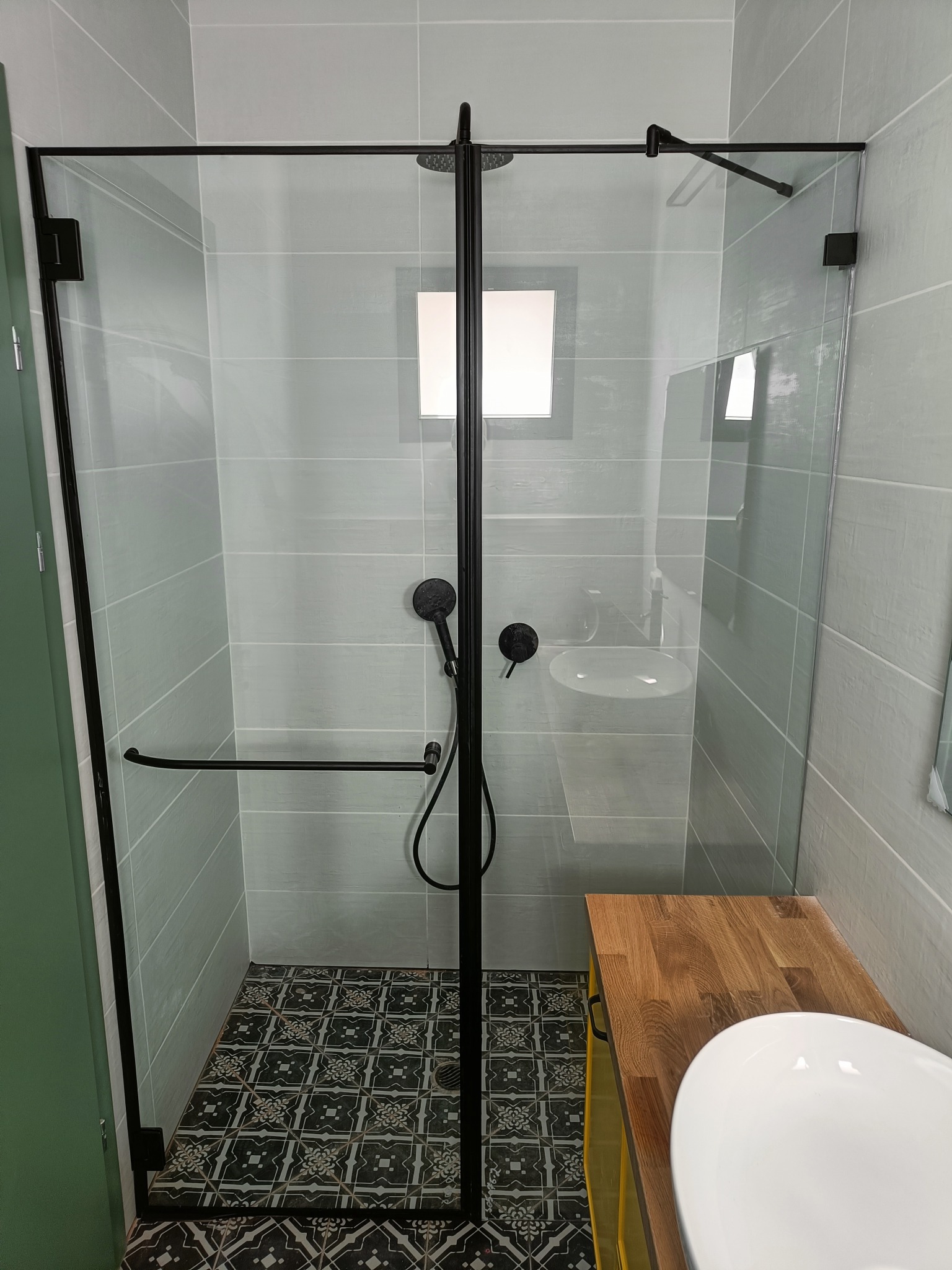 Kabina prysznicowa ze szklanymi drzwiami w nowoczesnej łazience z czarną armaturą i wzorzystą podłogą.