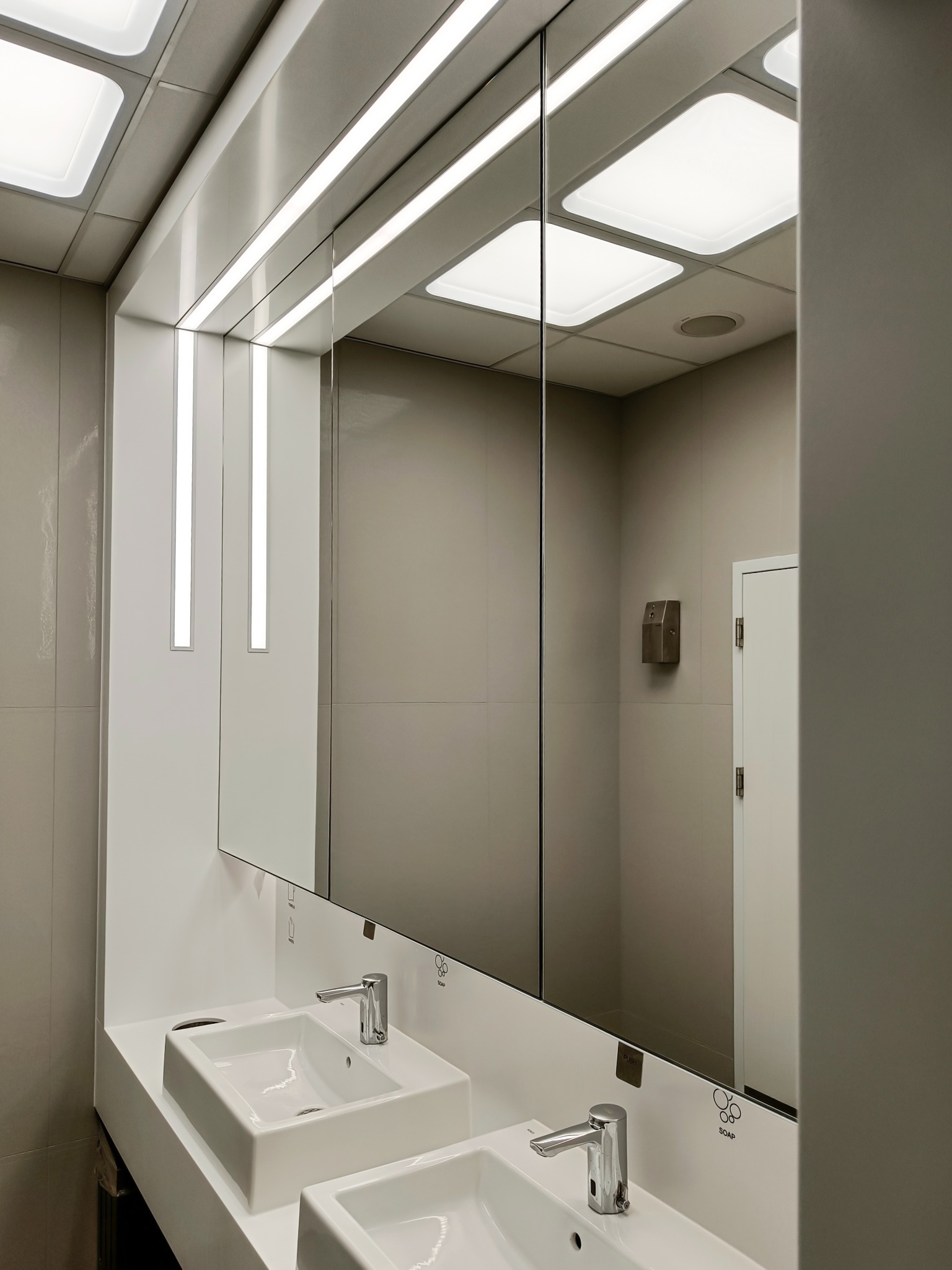 Nowoczesna łazienka biurowa z dwiema białymi umywalkami, dużym lustrem z podświetleniem LED i dozownikiem mydła na ścianie z szarych płytek.